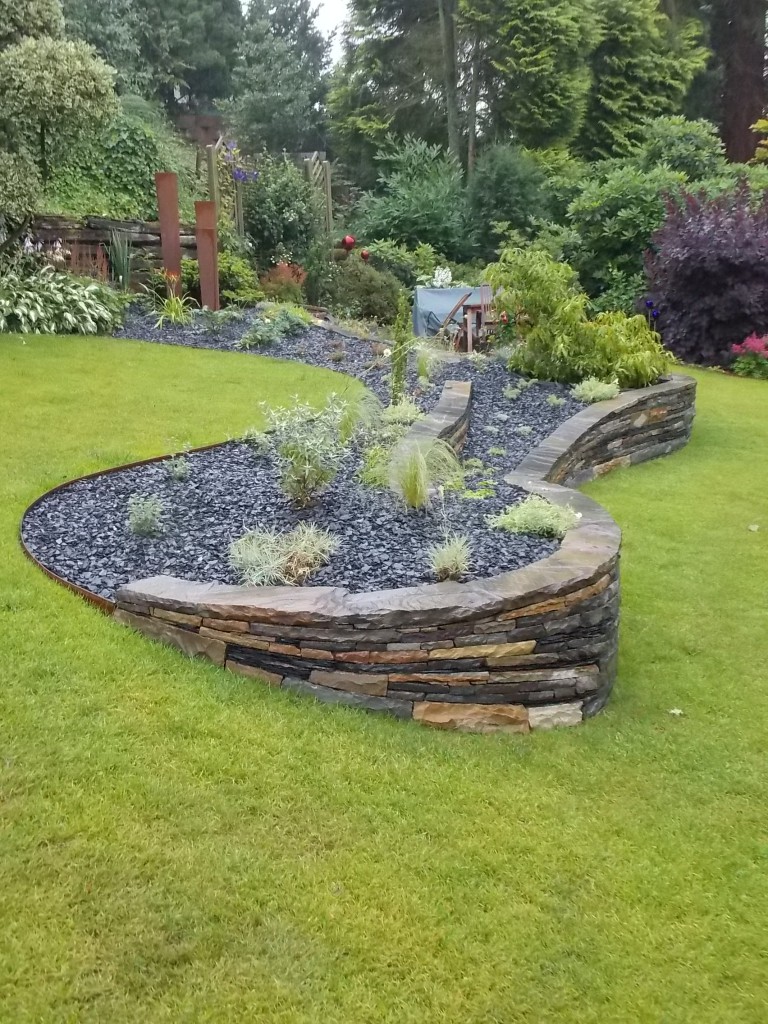 Die zugehörigen Flächen sind mit graulaubigen , niedrigwachsenden Stauden Bepflanzt. Die Fläche ist mit Schiefersplitt abgedeckt. Im Hintergrund ein Brunnen aus corten Stahl.